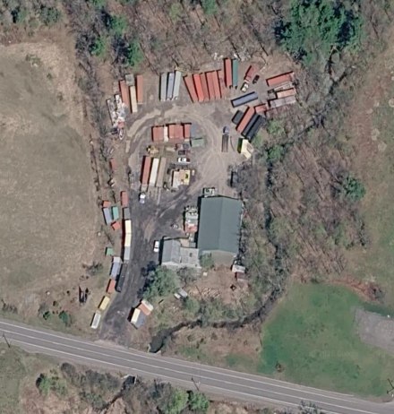 Satellite view of the C&C yard on Route 145 in East Durham: rows of shipping containers around a green-roofed workshop beside the road