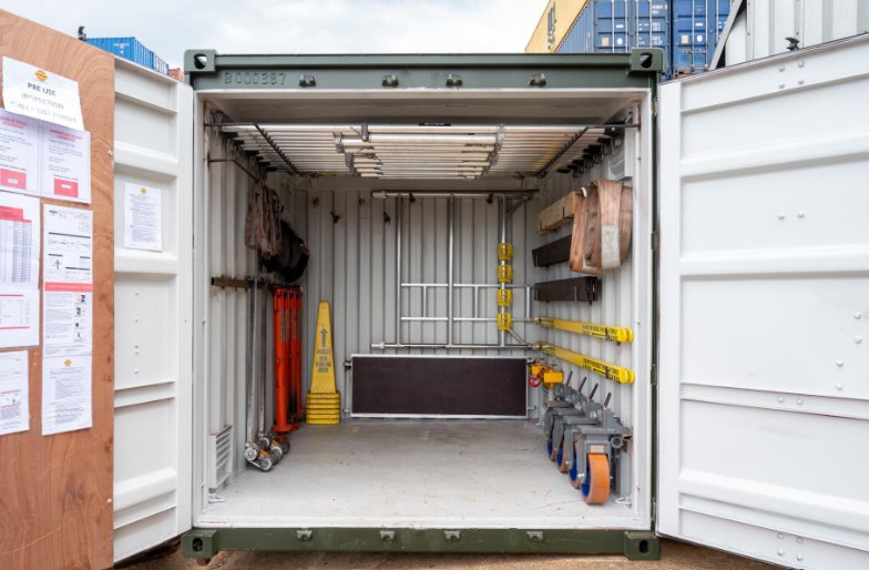An open shipping container fitted with ceiling racks, wall-mounted hooks, lifting straps, and wheeled equipment for tool storage